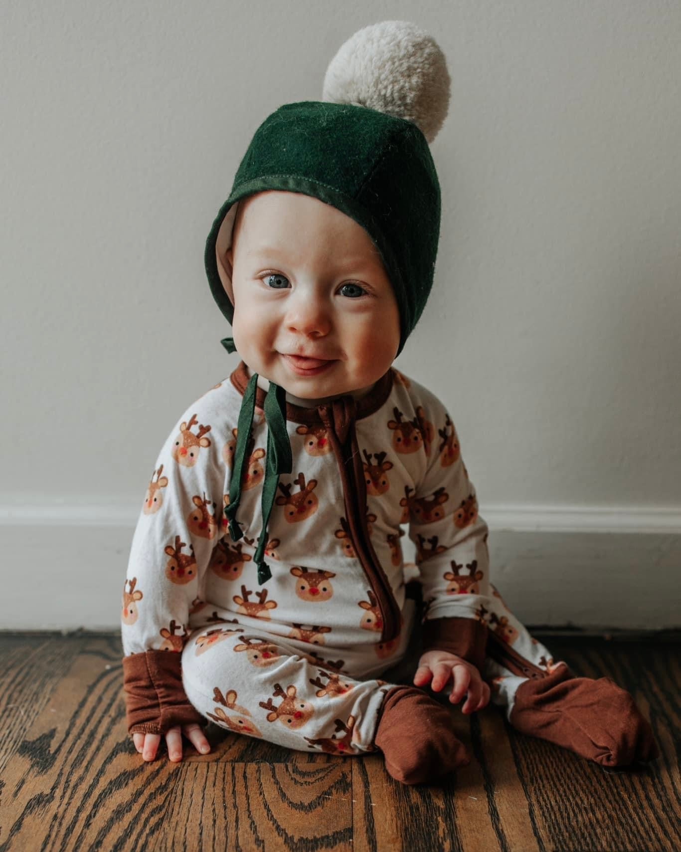 A baby in our Rudolph • Bamboo Zippy (Updated Fit), featuring a cream reindeer print, smiles at the camera while sitting on a wooden floor wearing a green hat with a white pom-pom.