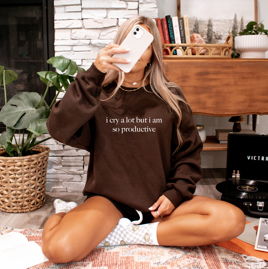 A woman sits cross-legged on the floor, holding her phone. Shes wearing the i cry a lot • Cocoa Pullover, a comfy brown cotton-poly blend sweatshirt. Books and plants surround her.