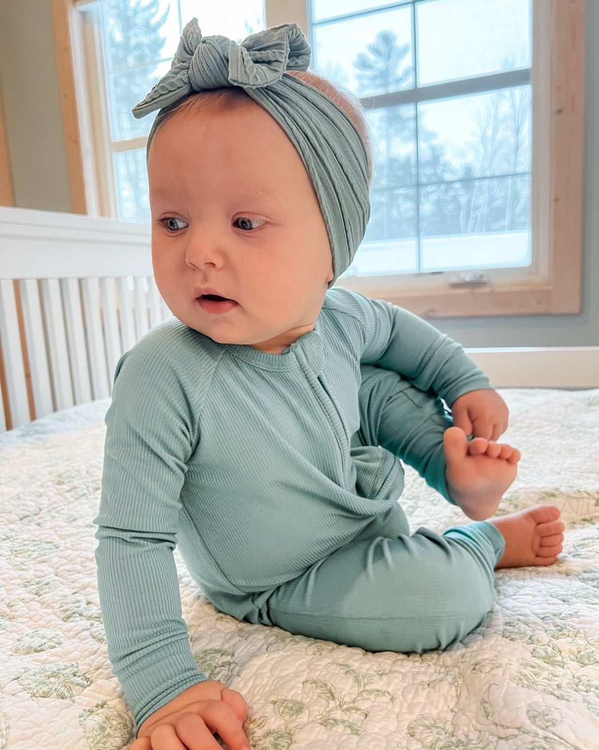 A baby in the Dusty Teal Ribbed Solid • Bamboo Zippy (Updated Fit) sits on a patterned quilt, glancing sideways. The cozy scene features a matching headband, white crib, and snowy outdoor view through large windows.