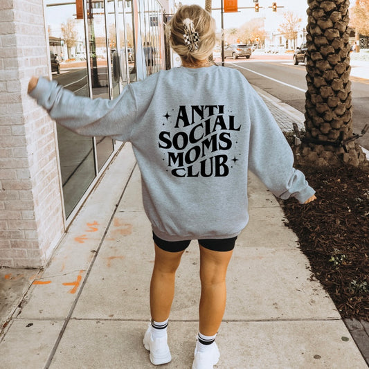 A woman with blonde hair in a messy bun walks on the sidewalk, wearing the Anti-Social Moms • Heather Gray Crew with “ANTI SOCIAL MOMS CLUB” in bold black letters on the back, paired with black shorts and white sneakers.