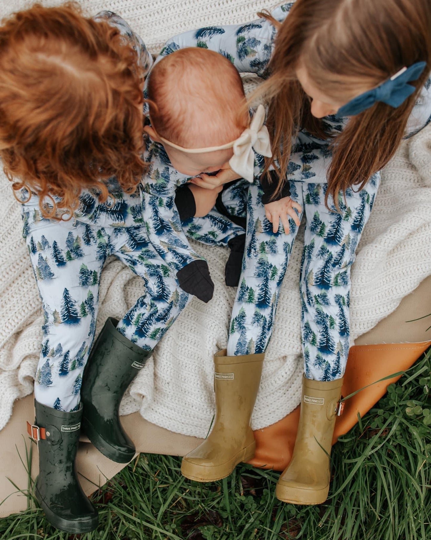 Three children sit on soft grass wearing matching Snowy Trees • 2 Piece Bamboo Set pajamas. Two older kids have rain boots, while the youngest baby with a bow headband sits between them. The eco-friendly bamboo fabric is gentle on sensitive skin.