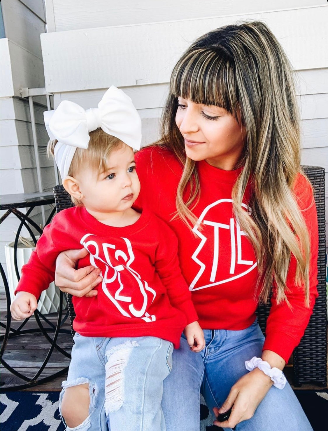 A woman and her bow-adorned toddler sit on a porch bench, both smiling in matching red Bestie Pullover • Red tops with white broken heart designs, showing off the perfect unisex youth fit alongside the womens style.
