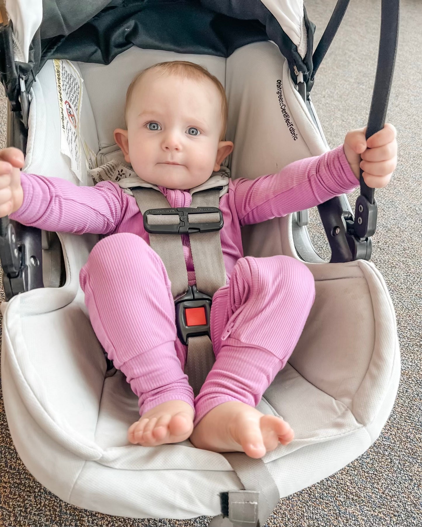 A baby wears the Lilac Ribbed Solid • Bamboo Zippy (Updated Fit), sitting barefoot in a car seat. The bamboo fabric is gentle on eczema-prone skin, perfect for comfort indoors on a carpeted floor.