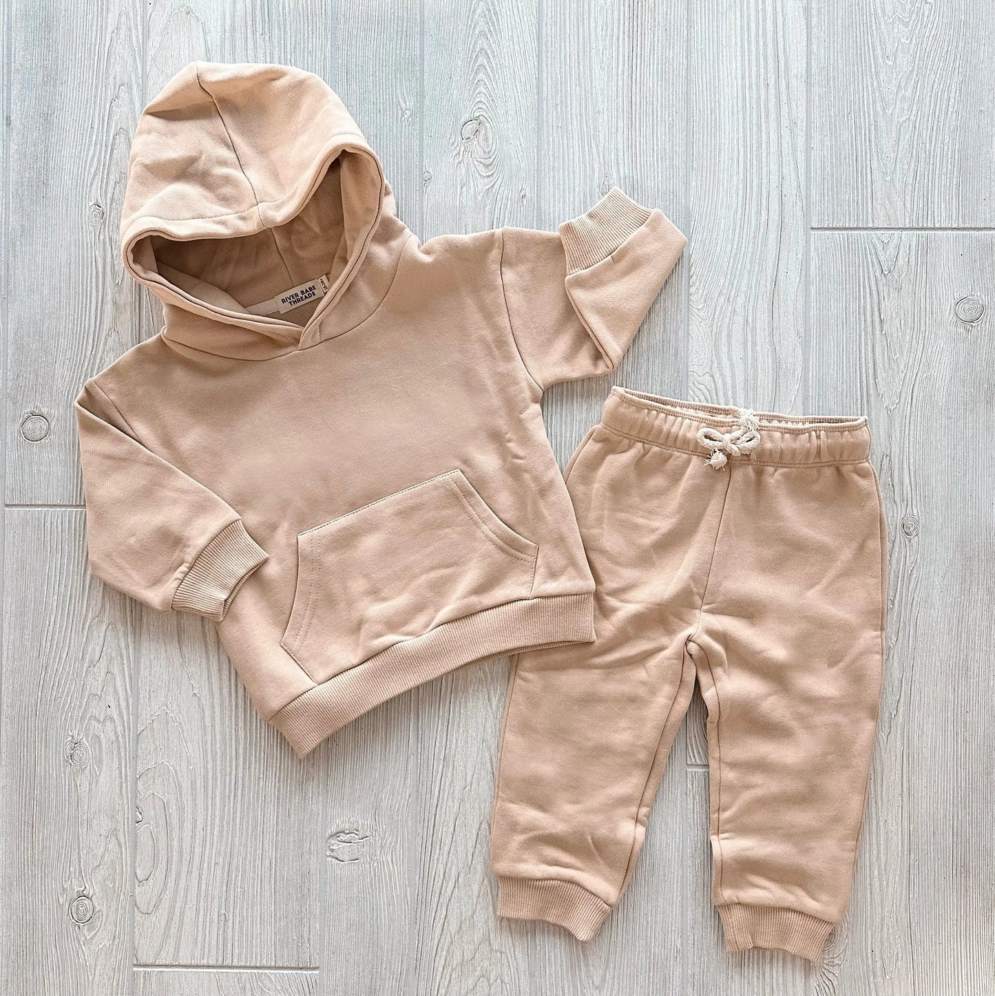 The Sand • Sweatpant & Hoodie Set is an organic beige two-piece for toddlers or babies, with a cozy fleece hoodie and adjustable drawstring sweatpants, both shown flat on a light wood floor.