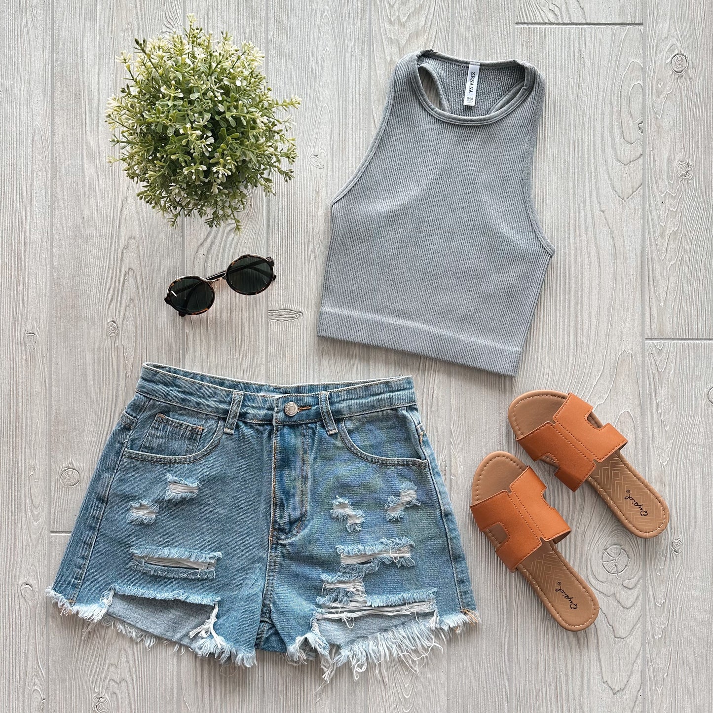 A flat lay of a casual summer outfit featuring the Sophia High-Neck Crop in Ash Gray, distressed denim shorts, tan slide sandals, black round sunglasses, and a small potted plant on a light wood floor.