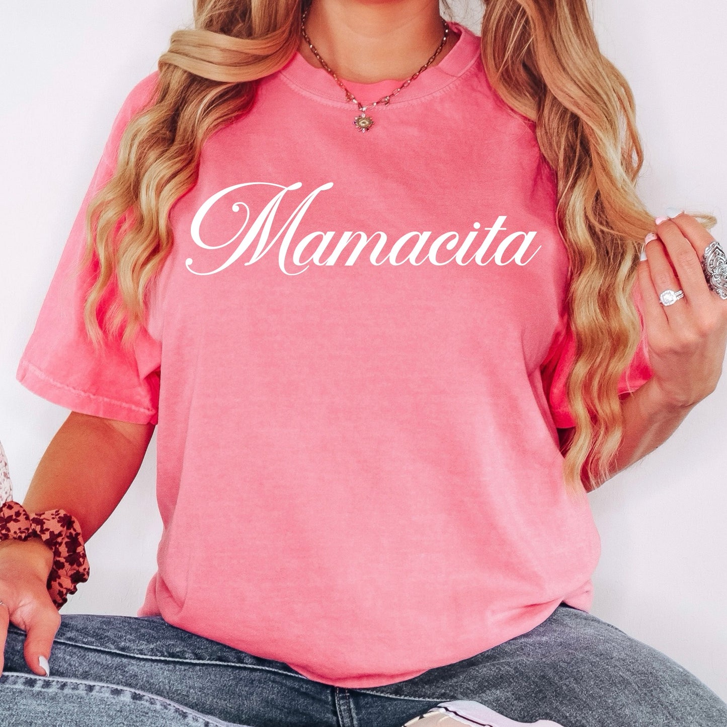 A woman sits casually in the New Mamacita • Garment Dyed Tee, her wavy hair, jewelry, and blue jeans completing her relaxed look.