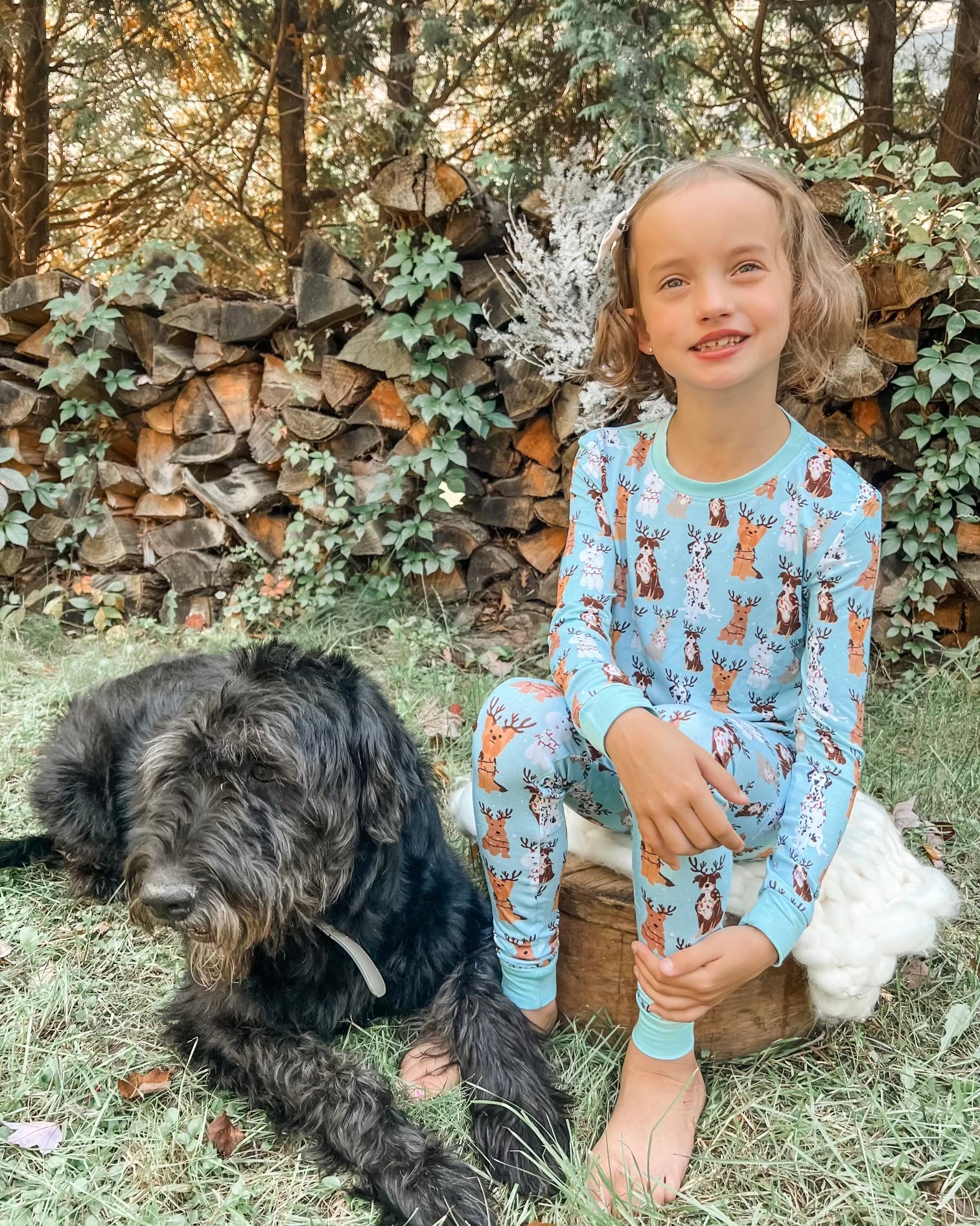 A young girl wears the Festive Friends • 2 Piece Bamboo Set (Updated Fit)—blue pajamas with reindeer prints—as she smiles on a tree stump outdoors next to a large black dog, with a woodpile and green vines in the background.