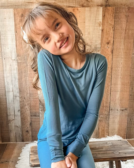 A young girl with light brown hair and a white bow smiles, wearing the Deep Blue Ribbed Solid • 2 Piece Bamboo Set (Updated Fit)—a soft, stretchy long-sleeve outfit ideal for sensitive skin—as she sits on a rustic wooden bench.