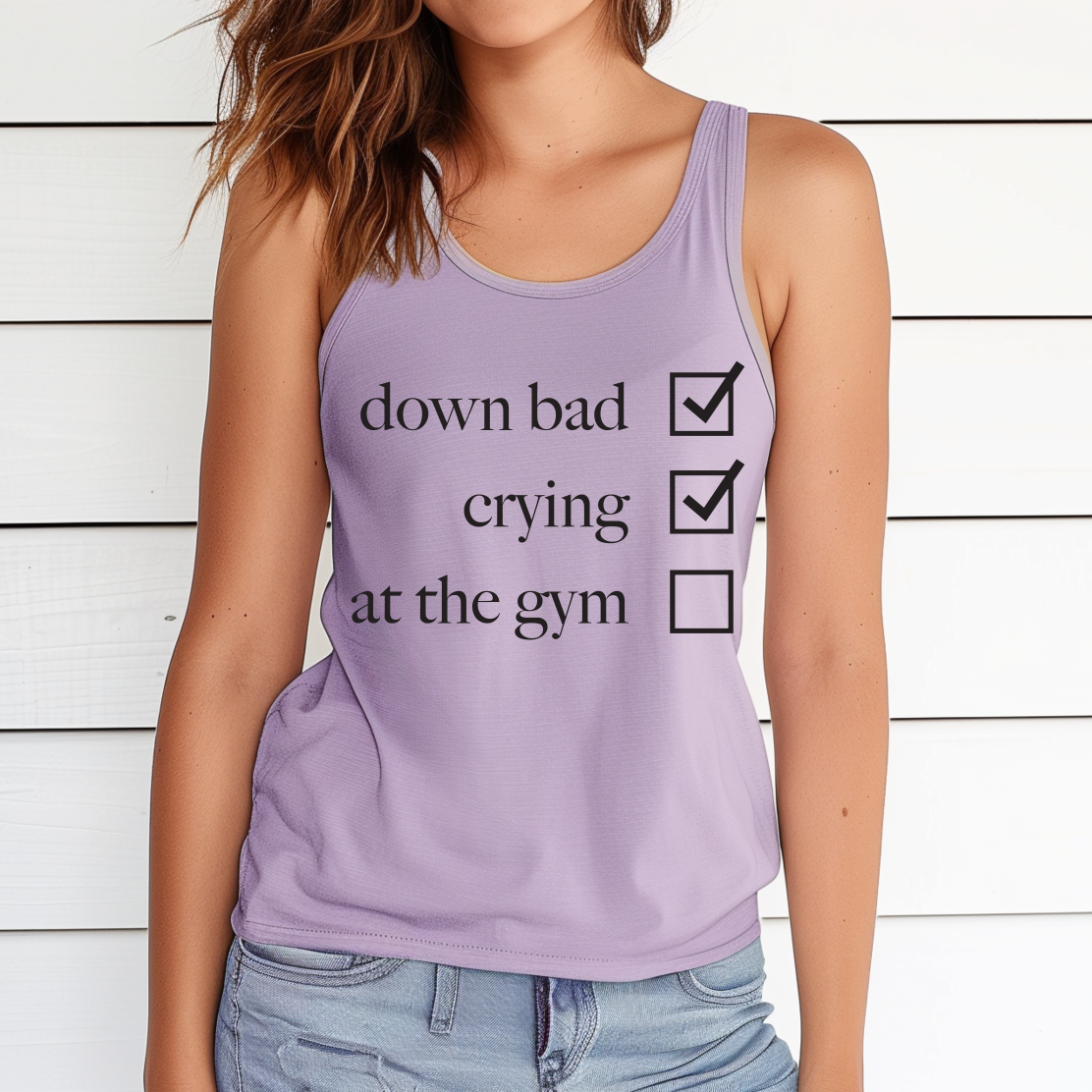 A woman models the Not at the Gym • Lilac Tank—ideal for summer—featuring down bad and crying checked, at the gym unchecked. She poses by a white wooden wall, highlighting this chic womens fit.