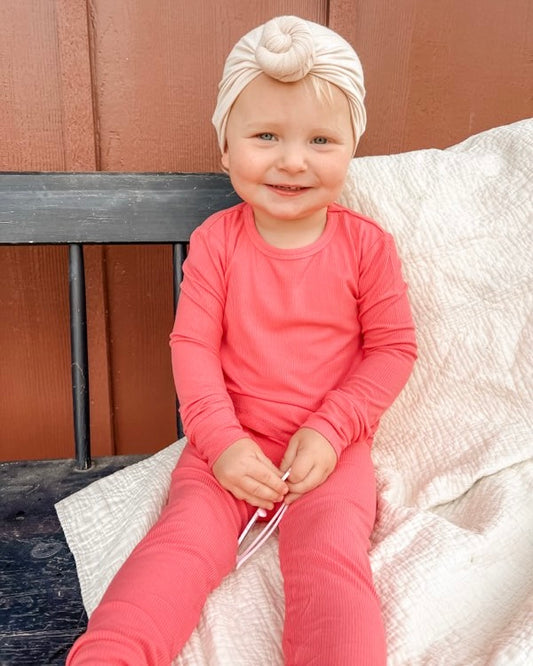 A smiling toddler in the Bright Pink Ribbed Solid • 2 Piece Bamboo Set (Updated Fit) sits on a bench with a beige knotted headband and a white textured blanket, in front of a wooden wall.