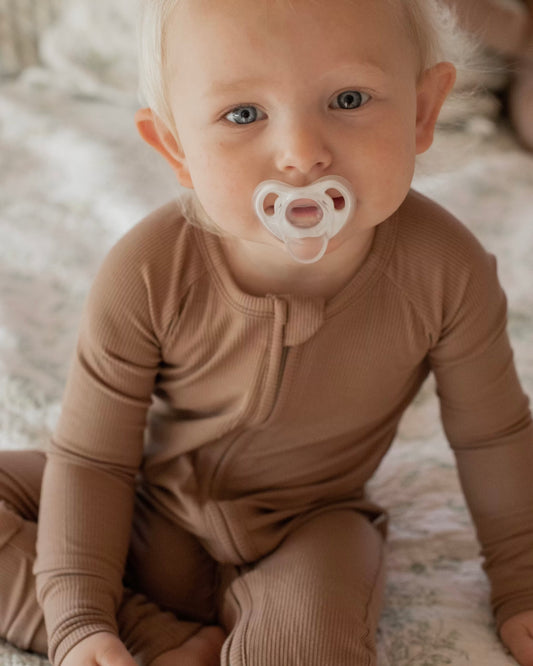 A young child with blond hair and blue eyes sits on a bed, wearing the Chestnut Ribbed Solid • Bamboo Zippy (Updated Fit) and holding a white pacifier in their mouth.