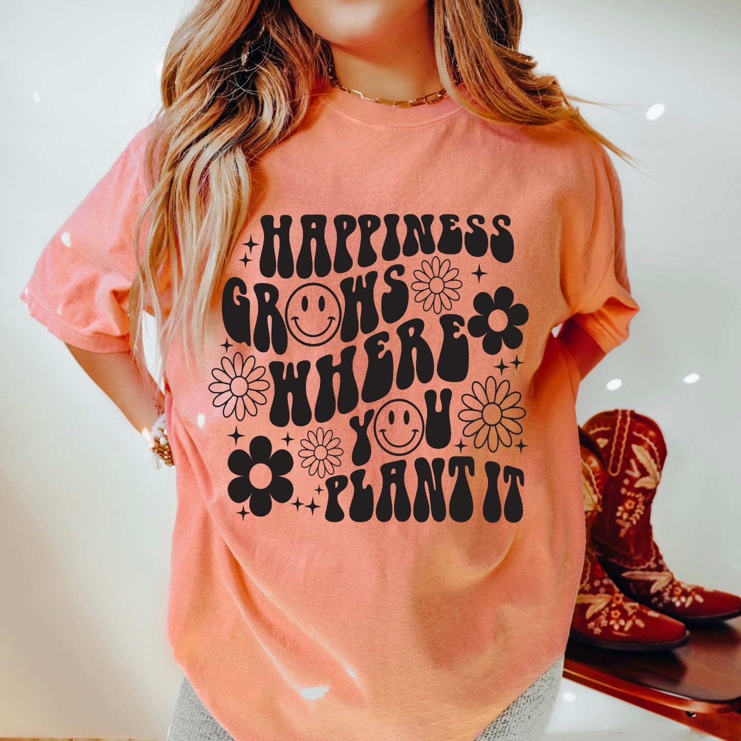 A woman wears the Happiness Grows • Comfort Colors Tee, a peach ring-spun cotton shirt featuring black retro text, flower designs, smiley faces, and the phrase Happiness grows where you plant it.