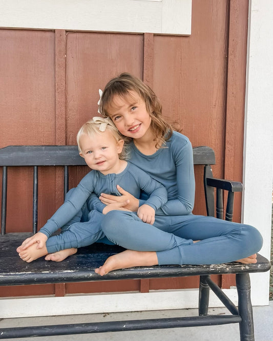 Two young girls wear matching Deep Blue Ribbed Solid • 2 Piece Bamboo Sets with updated fit as they sit on a black bench in front of a red and white wooden wall. The older girl hugs the younger one, both smiling at the camera.