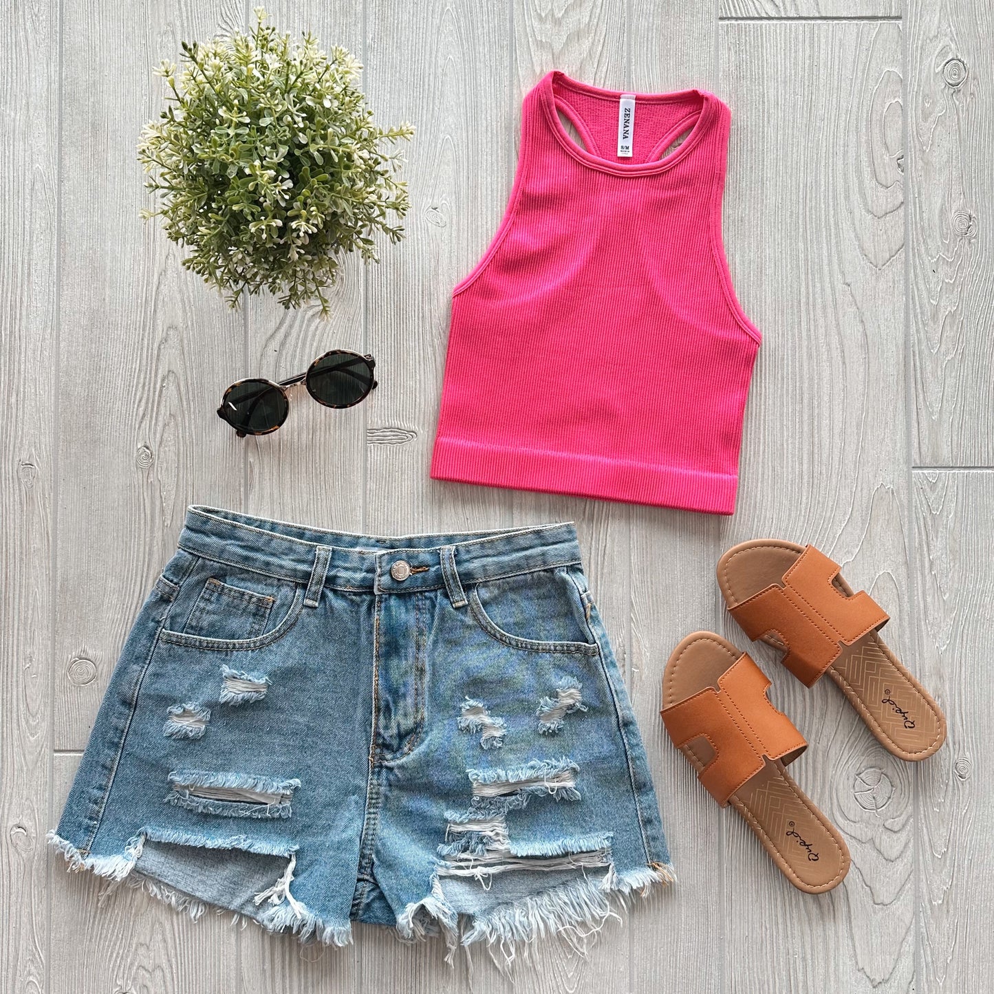 Flat lay of a summer outfit featuring the Sophia High-Neck Crop in pink, distressed denim shorts, tan slip-on sandals, round black sunglasses, and a small green potted plant on a light wooden floor.