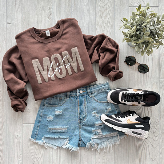 Flat lay of a Mom Life • Chocolate Tonal sweatshirt, distressed denim shorts, black and white Nike sneakers, round black sunglasses, and a small potted plant on a light wooden floor.
