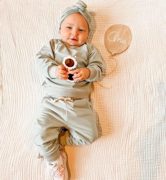 A baby in the Mint • Sweatpant & Crew Set with adjustable drawstring pants and matching headband lies on a cream quilt, holding a teething toy. A wooden One plaque marks their first birthday.