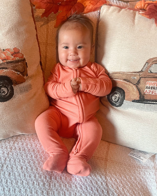 A smiling baby in the Coral Ribbed Solid • Bamboo Zippy (Updated Fit) onesie sits on a couch between two fall-themed pillows with vintage truck and pumpkin designs.
