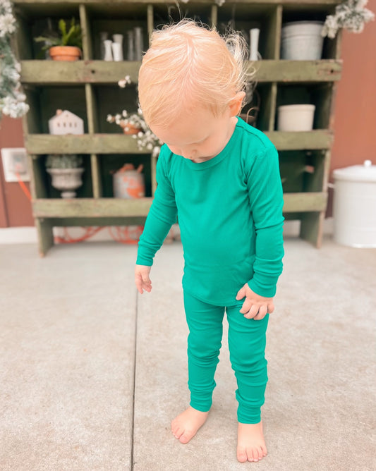 A young blonde child, barefoot on a concrete floor, wears the Emerald Ribbed Solid • 2 Piece Bamboo Set (Updated Fit) in bright green. Behind them is a green wooden shelf with various items as they look down.