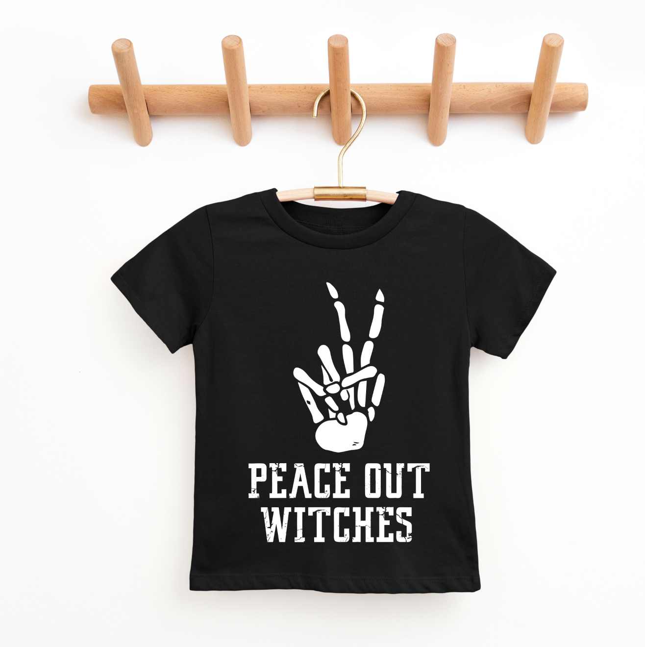The Peace Out Witches Kids Tee is a black, true-to-size shirt featuring a white skeleton hand peace sign and PEACE OUT WITCHES text, shown on a wooden rack—perfect for unisex kids when shopping small.