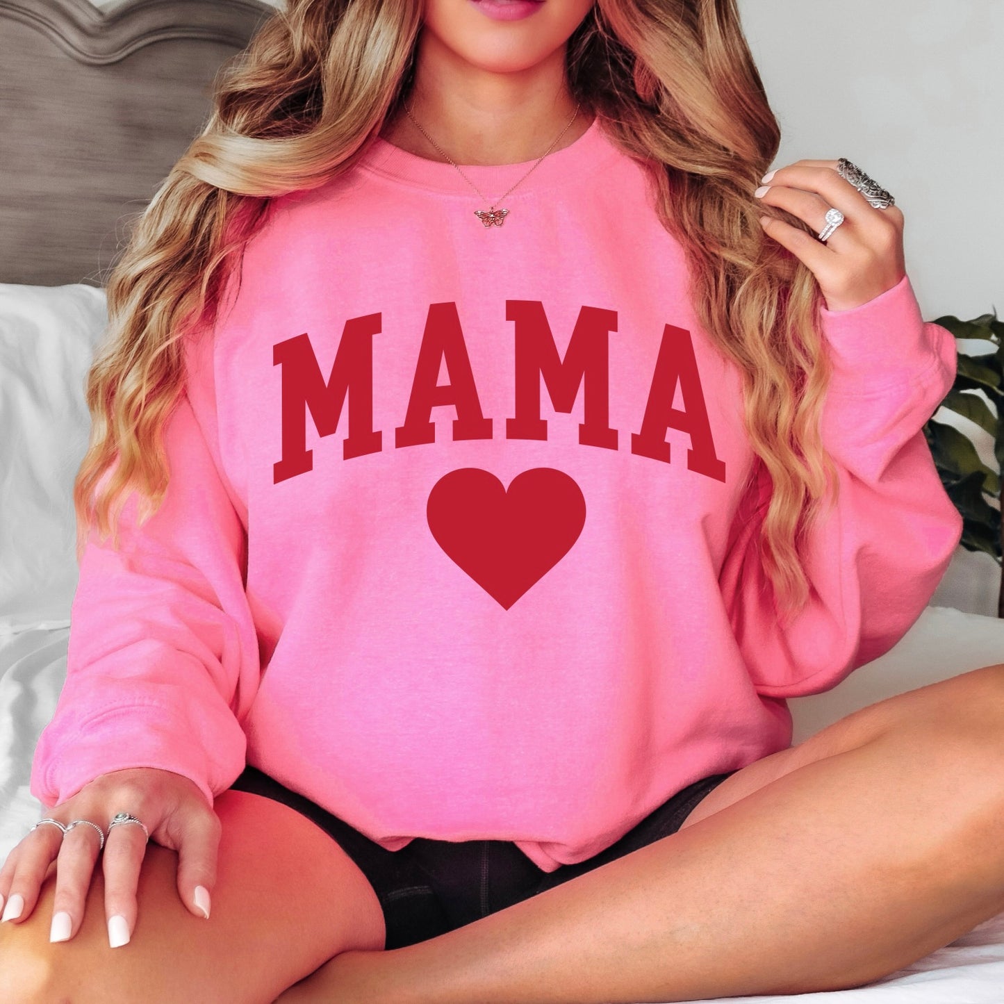 A woman with long wavy hair sits on a bed wearing the Mama Love • Neon Pink Pullover, featuring a bold red “MAMA” and heart design. She showcases the cozy unisex fit of this stylish Valentine’s Day sweatshirt.