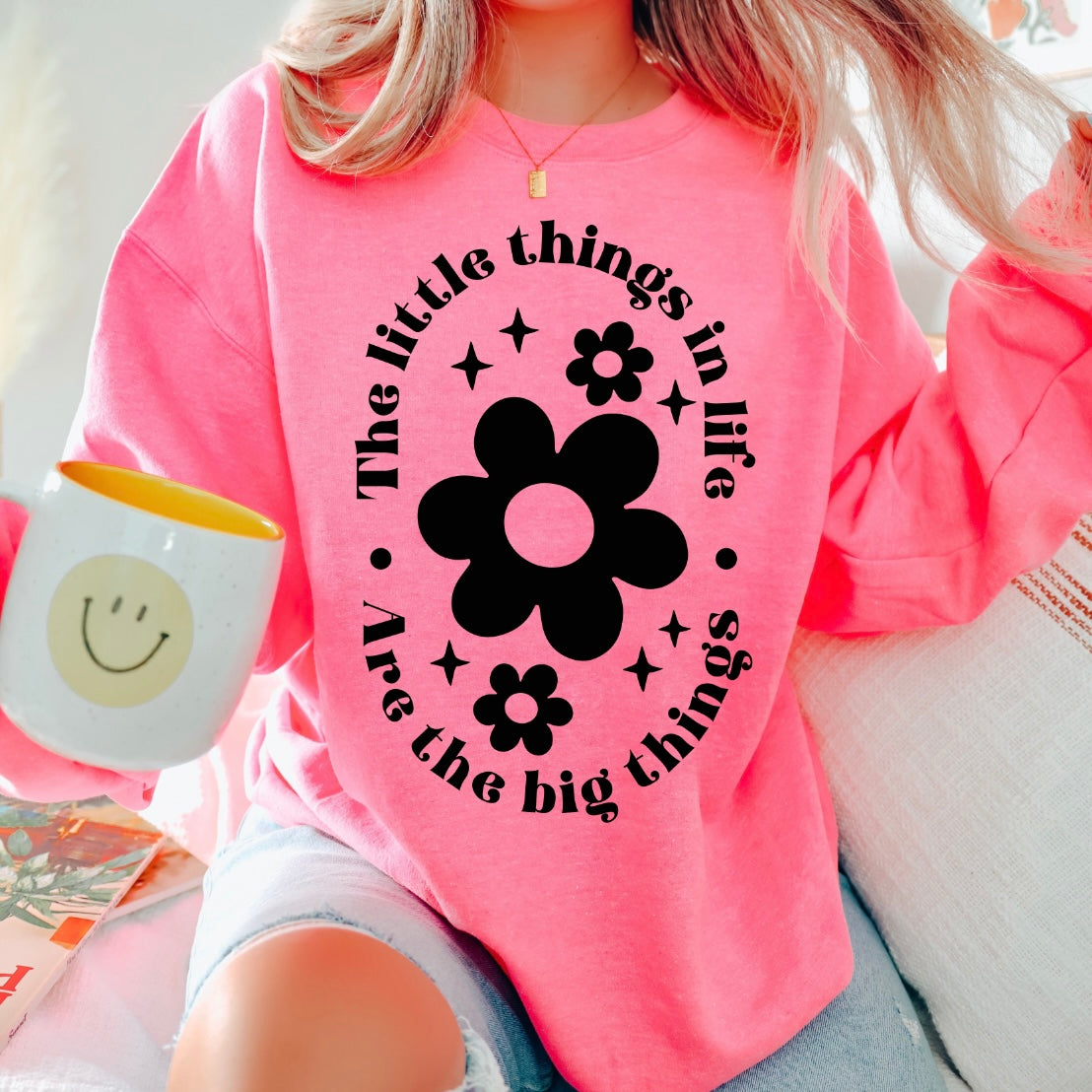 Wearing the Neon Pink The Little Things Pullover, featuring black flower graphics and the phrase “The little things in life are the big things,” a person holds a smiley face mug, radiating cozy, cheerful energy.