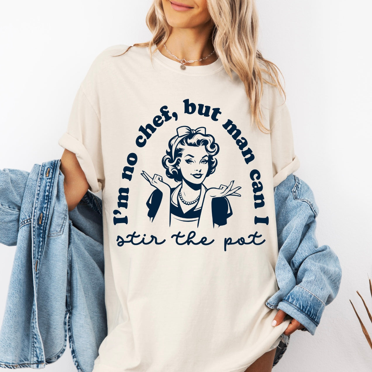 A woman wears the Stir the Pot • Comfort Colors Tee, a loose beige shirt made from soft ring-spun cotton with a retro-style illustration and the text: I’m no chef, but man can I stir the pot. She has a denim jacket draped over her arms.