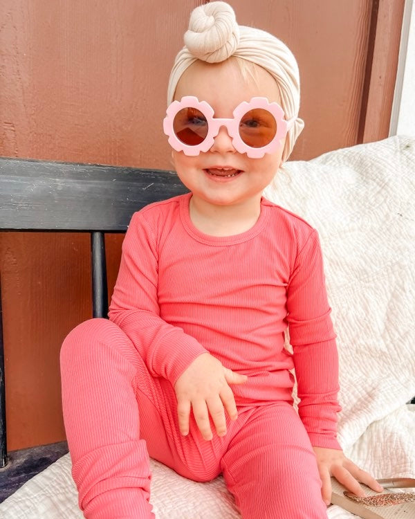 A smiling toddler in our Bright Pink Ribbed Solid 2 Piece Bamboo Set (Updated Fit), pink flower sunglasses, and a beige headwrap sits on a bench against a brown wall.