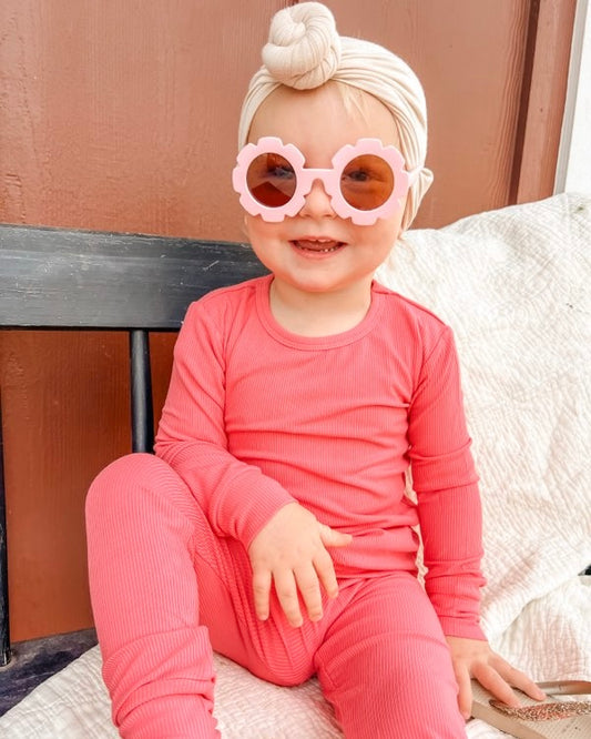 A smiling toddler in our Bright Pink Ribbed Solid 2 Piece Bamboo Set (Updated Fit), pink flower sunglasses, and a beige headwrap sits on a bench against a brown wall.