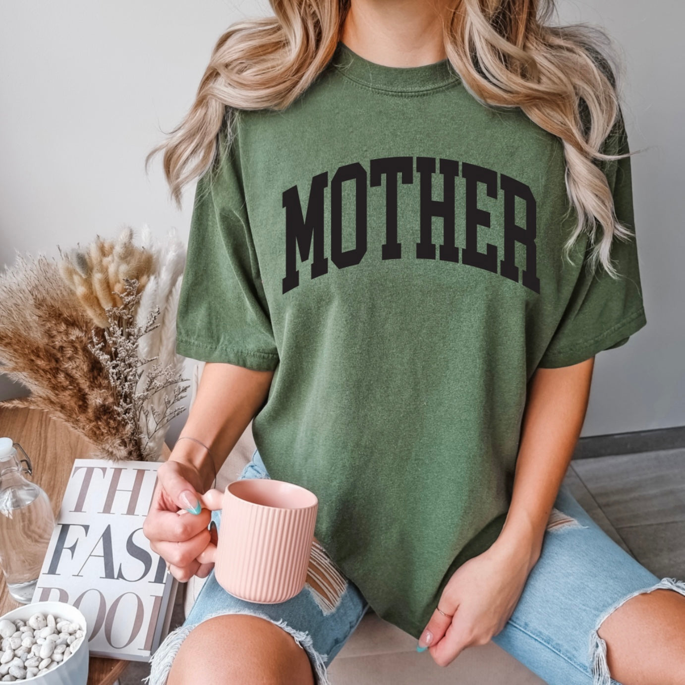 A woman with wavy blonde hair and ripped blue jeans sits on a couch holding a pink mug, wearing the MOTHER • Monterey Sage Dyed Tee. She is surrounded by dried flowers and books.