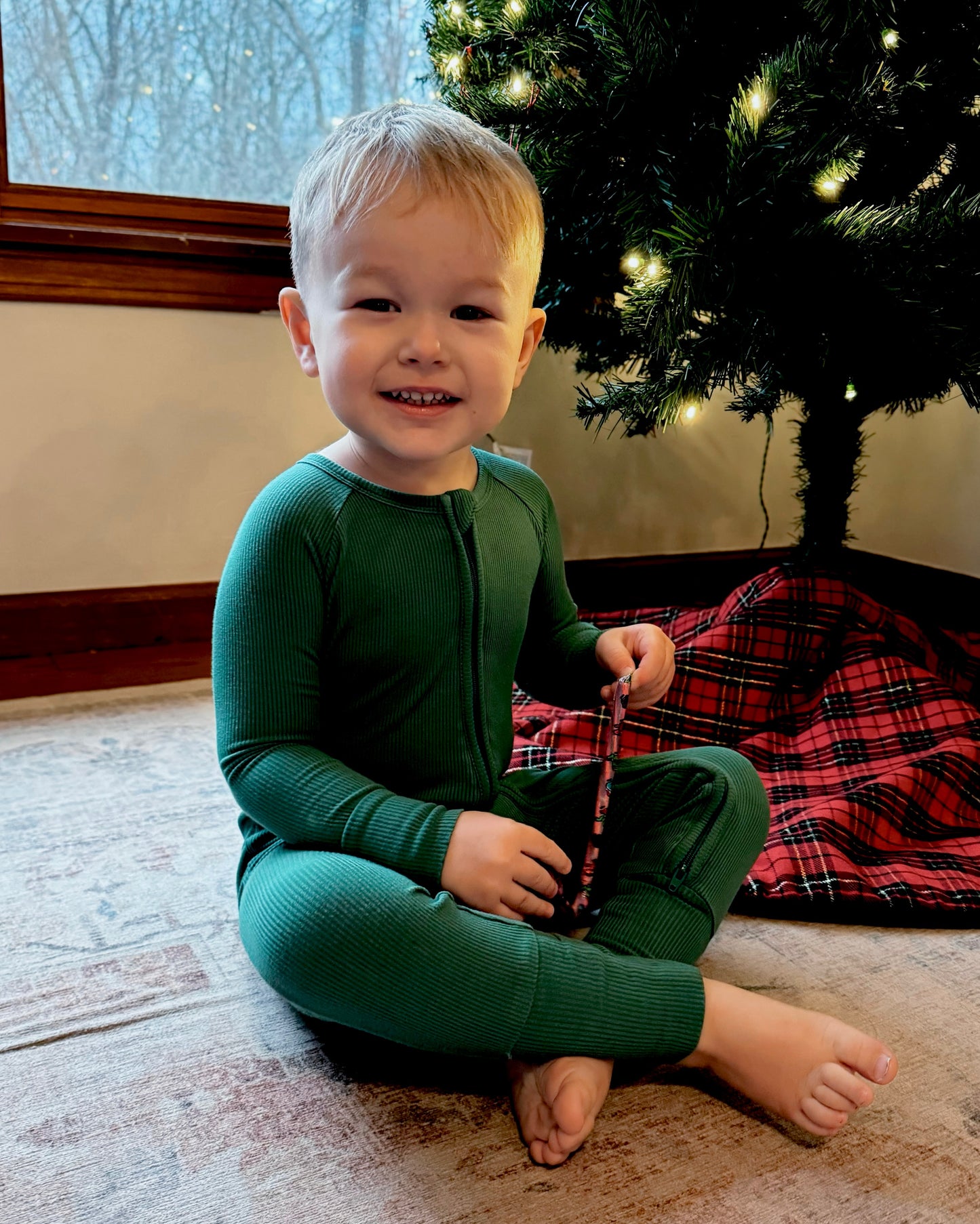 A smiling child in Forest Green Ribbed Solid • Bamboo Zippy (Updated Fit) pajamas sits by a Christmas tree with lights, holding a candy cane. A red plaid tree skirt is partially visible beneath the tree.