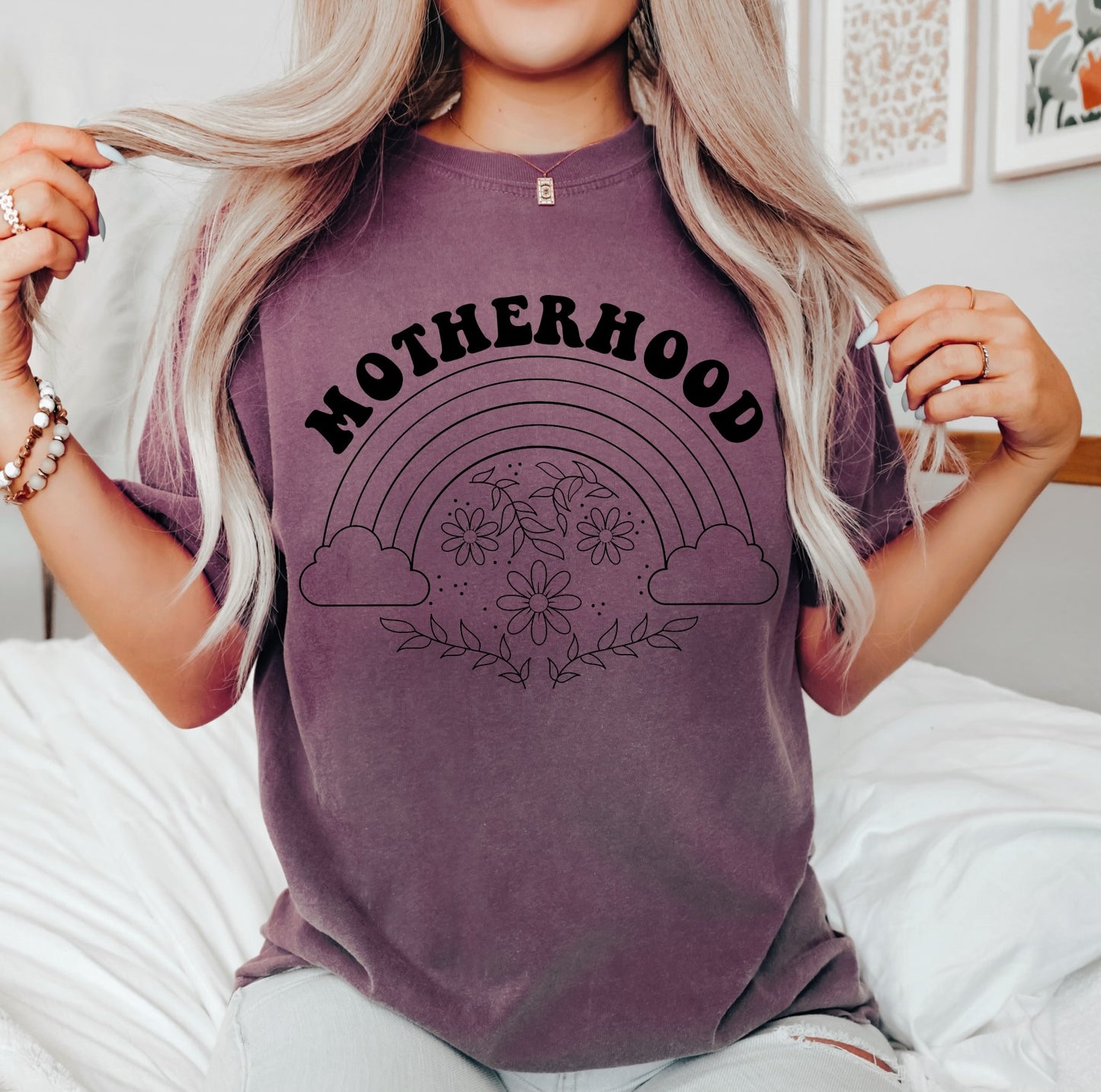 A woman with long blonde hair sits on a bed wearing the Motherhood • Vineyard Dyed Tee, which features MOTHERHOOD above a rainbow, clouds, and floral design. She holds her hair and wears beaded bracelets, with framed art behind her.