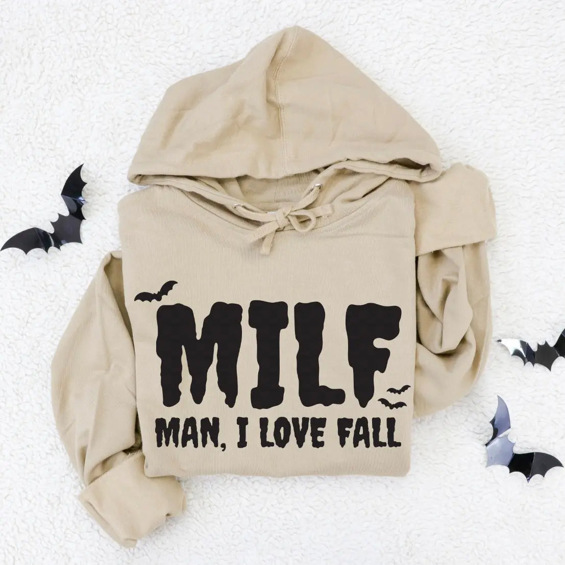 The Spooky MILF • Tan Hoodie features MILF MAN, I LOVE FALL in bold black letters with bat illustrations. This comfy unisex hoodie is perfect for autumn and is shown laid out on a white textured surface with black paper bats.