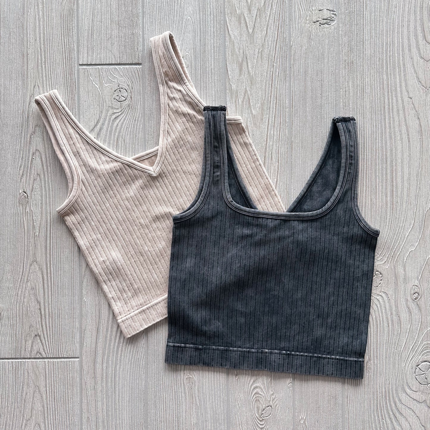 Two Ava Ribbed Crops from the Vintage Collection are laid flat on a light wooden surface—one beige, one dark gray overlapping it. Both feature wide straps, a V-neckline, and are made of soft, stretchy fabric.