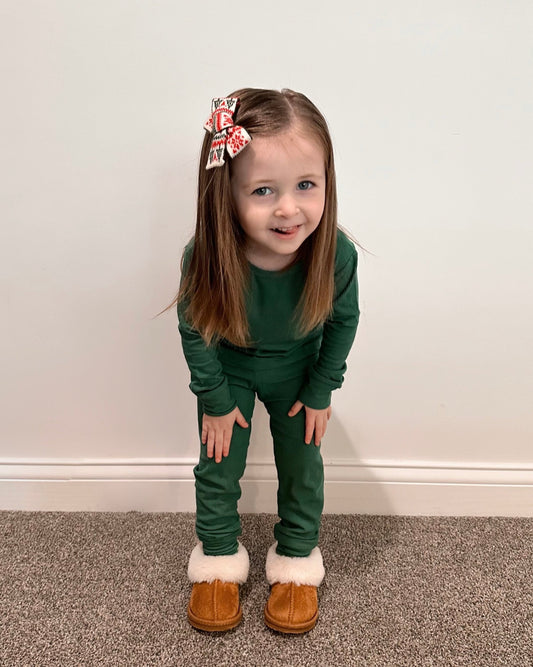 A young girl with long brown hair and a large festive bow wears the Forest Green Ribbed Solid • 2 Piece Bamboo Set (Updated Fit) and brown slippers with white fur, standing indoors on a carpeted floor and smiling at the camera.