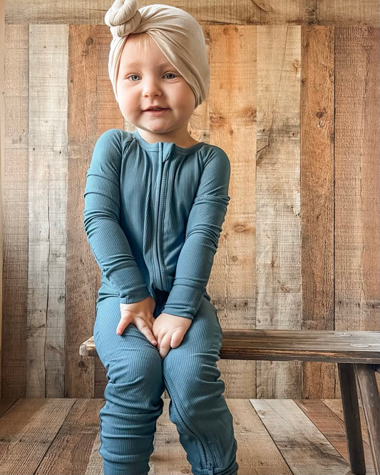 A young child in a light beige headwrap and Deep Blue Ribbed Solid • Bamboo Zippy (Updated Fit) sits on a rustic wooden bench, smiling gently—this soft jumpsuit is ideal for sensitive skin.