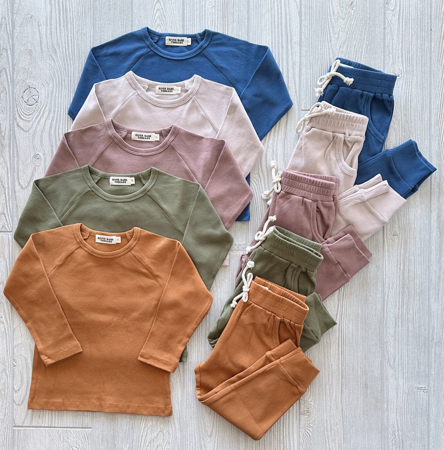 Five Ribbed Organic Cotton Jogger Sets in blue, light pink, olive green, mauve, and mustard are neatly arranged on a light wooden floor. Each set includes a matching long-sleeve top and drawstring jogger pants.