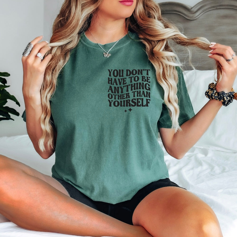A woman with long wavy hair sits cross-legged on a bed wearing the Self Love • Comfort Colors Tee—100% ring-spun cotton, green, with YOU DONT HAVE TO BE ANYTHING OTHER THAN YOURSELF—plus black shorts as she holds her hair.