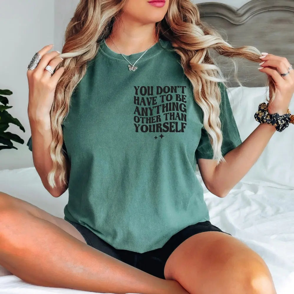 A woman with long wavy hair sits cross-legged on a bed wearing the Self Love • Comfort Colors Tee—100% ring-spun cotton, green, with YOU DONT HAVE TO BE ANYTHING OTHER THAN YOURSELF—plus black shorts as she holds her hair.