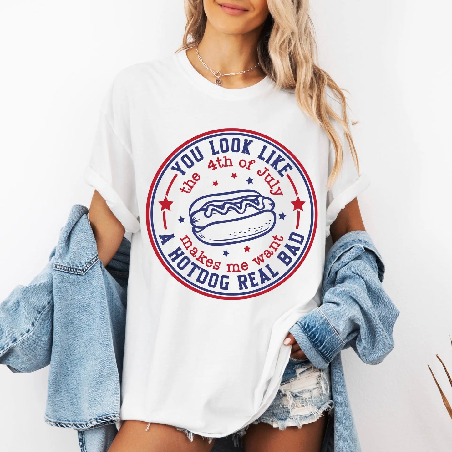 A woman with light hair wears the 4th of July • Comfort Colors Tee—a white shirt with a hot dog graphic and You look like the 4th of July, makes me want a hotdog real bad—plus denim shorts & a jacket draped over her arms.
