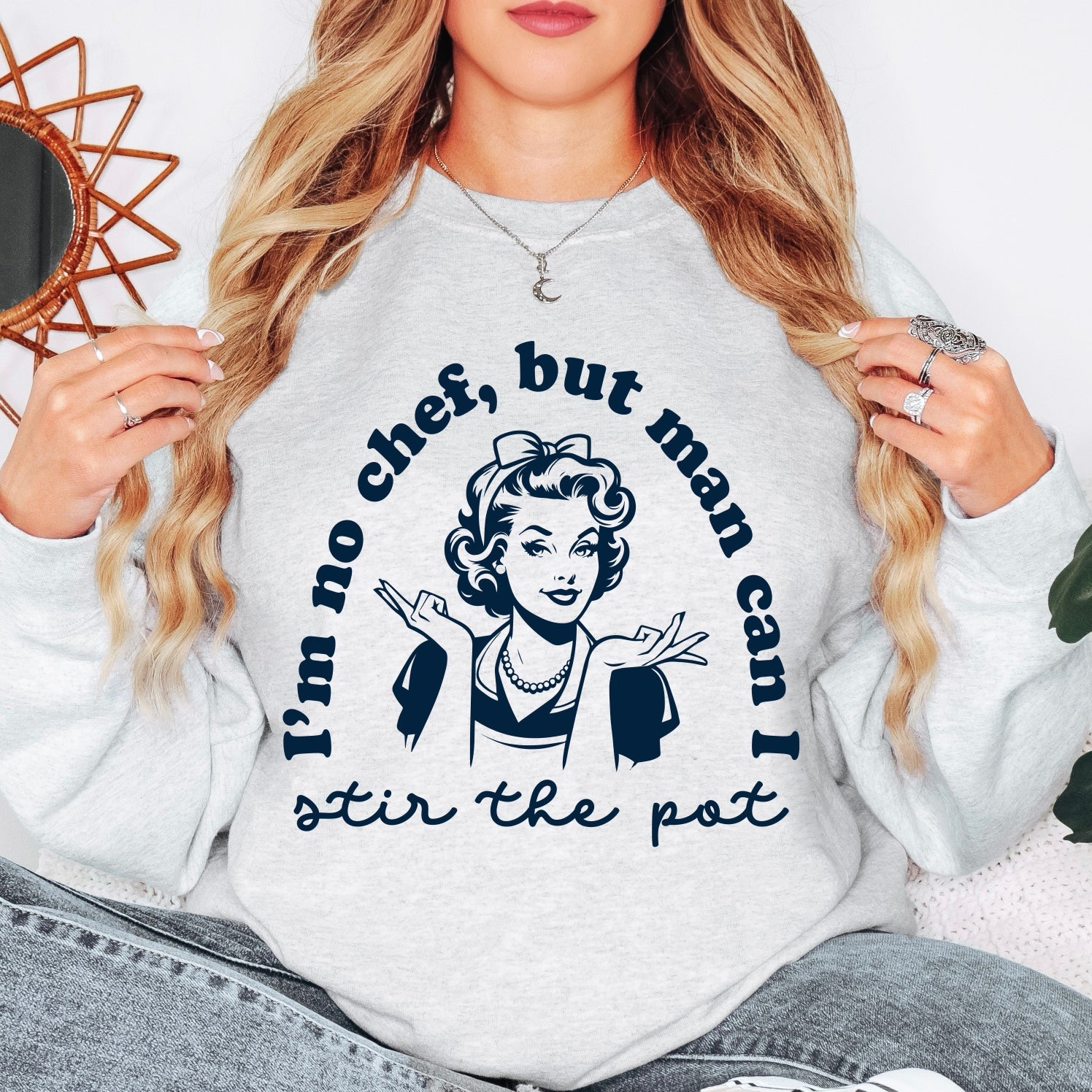 A person wears the Stir the Pot • Ash Pullover, a unisex gray sweatshirt with a retro woman graphic and the phrase Im no chef, but man can I stir the pot, enjoying its comfy fit with hands raised near their shoulders.