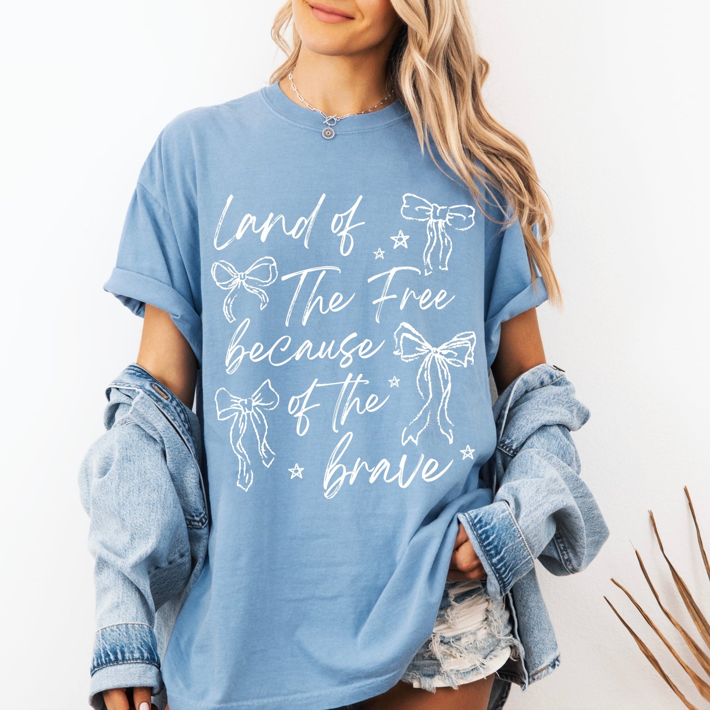 A woman with long blonde hair wears the Because of the Brave • Comfort Colors Tee featuring bow illustrations and the text Land of the Free because of the brave, styled with denim shorts and a jacket draped off her shoulders.