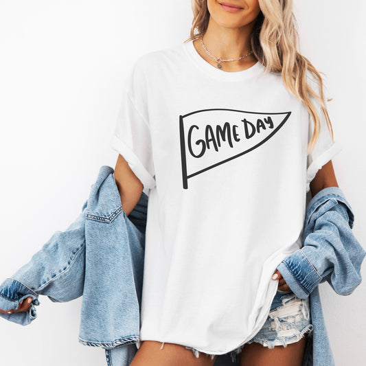 A woman wears the Game Day • Comfort Tee, a white garment-dyed shirt featuring a pennant graphic. Styled with a denim jacket and shorts, this tee is perfect for your fall wardrobe.