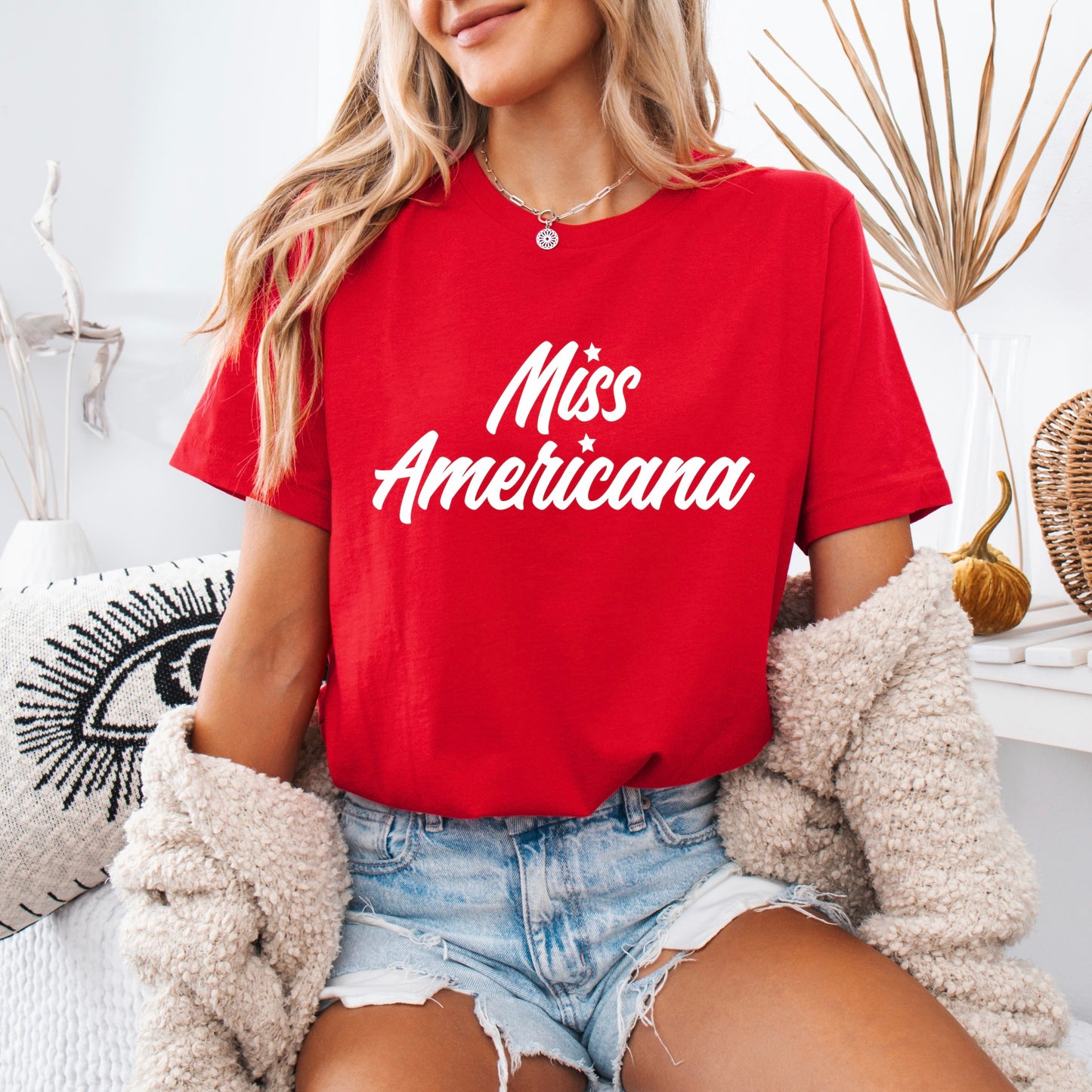 A woman wearing the Miss Americana • Tee, denim shorts, and a beige cardigan relaxes on a white couch with modern decor—a perfect look for any Swiftie fan.
