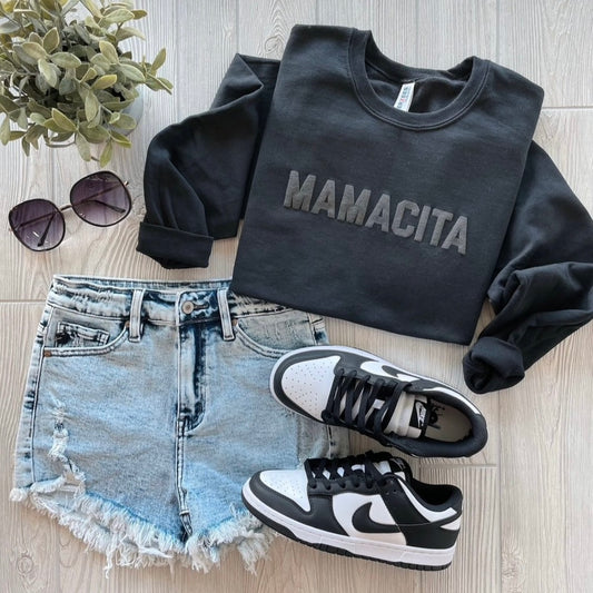 A flat lay of a black MAMACITA • Tonal Pullover, light blue distressed shorts, black and white Nike sneakers, sunglasses, and a green plant displayed on a light wood surface.
