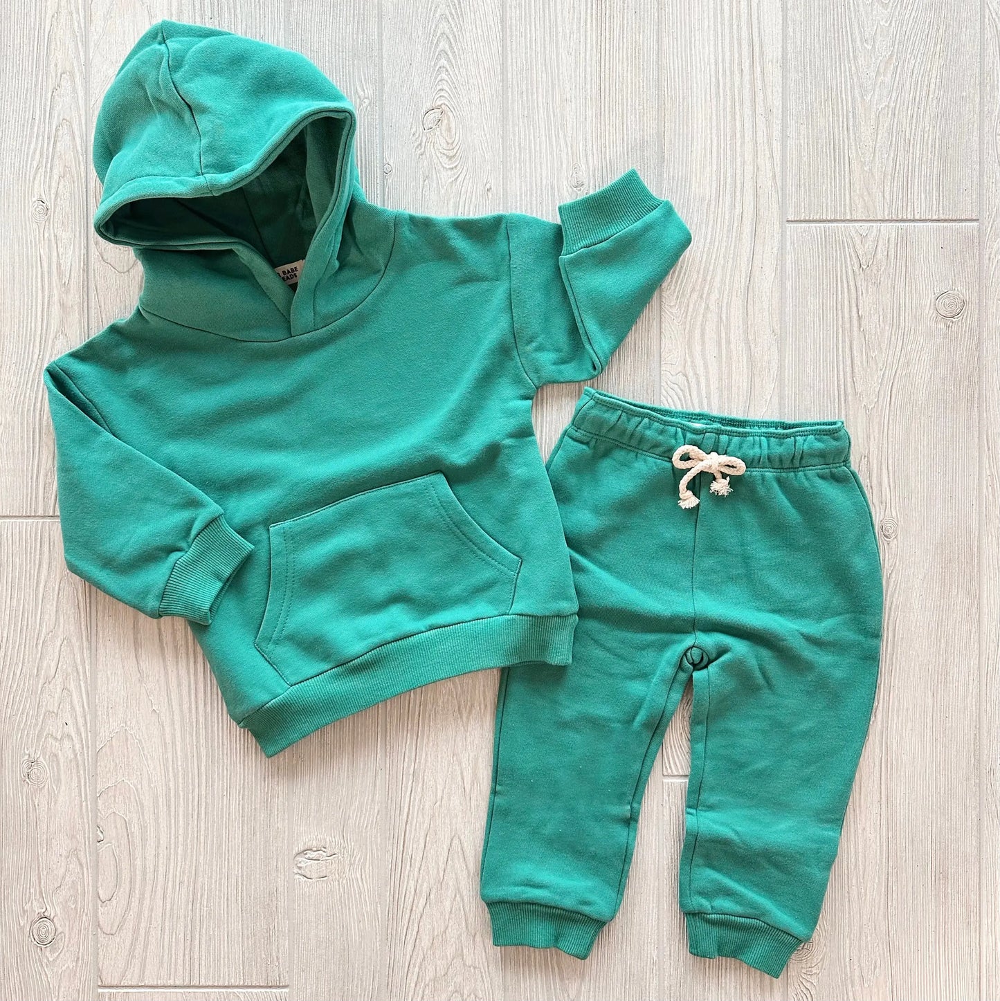 The Jade • Sweatpant & Hoodie Set is a green, toddler-sized organic two-piece featuring a fleece hoodie with front pocket and matching adjustable drawstring sweatpants, both displayed flat on a light wood surface.