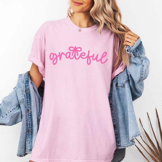 A woman wears the "grateful • Comfort Tee," a light pink, garment-dyed Comfort Colors T-shirt with “grateful” in pink script, paired with a light blue denim jacket—an easy fall wardrobe pick against a plain white background.