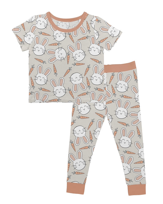 The Snuggle Bunny • 2 Piece Bamboo Set (Updated Fit) features a short-sleeve top and long pants with smiling bunny faces and carrots on a light beige background, accented with light brown cuffs and neckline.