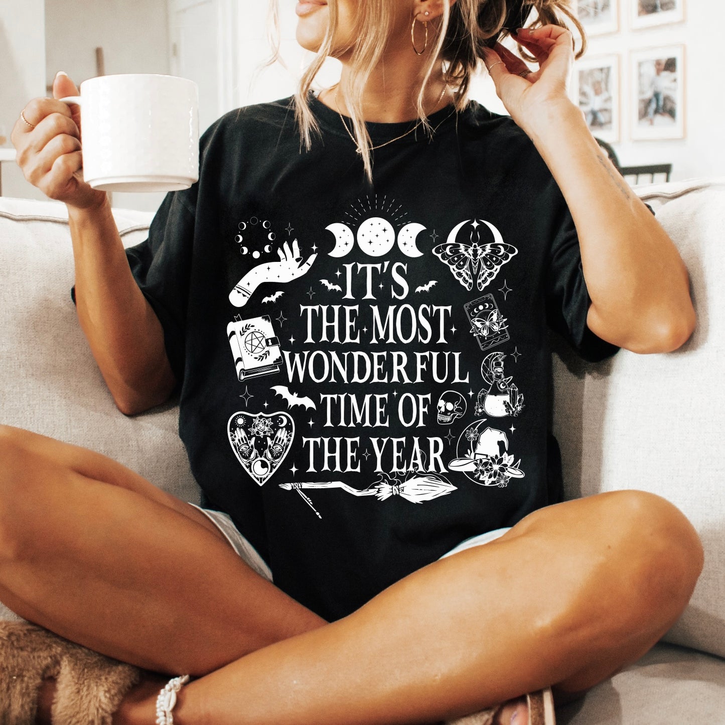 A woman sits on a couch holding a white mug, wearing the Most Wonderful Time • Comfort Tee—a black, soft ring-spun cotton shirt featuring mystical graphics and white text that says, Its the most wonderful time of the year.