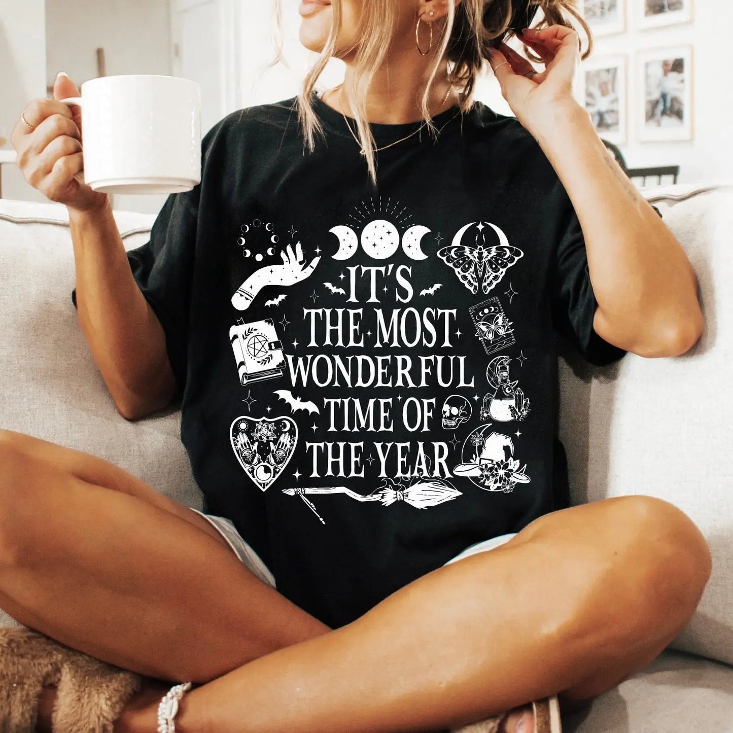 A woman sits on a couch holding a white mug, wearing the Most Wonderful Time • Comfort Tee—a black, soft ring-spun cotton shirt featuring mystical graphics and white text that says, Its the most wonderful time of the year.