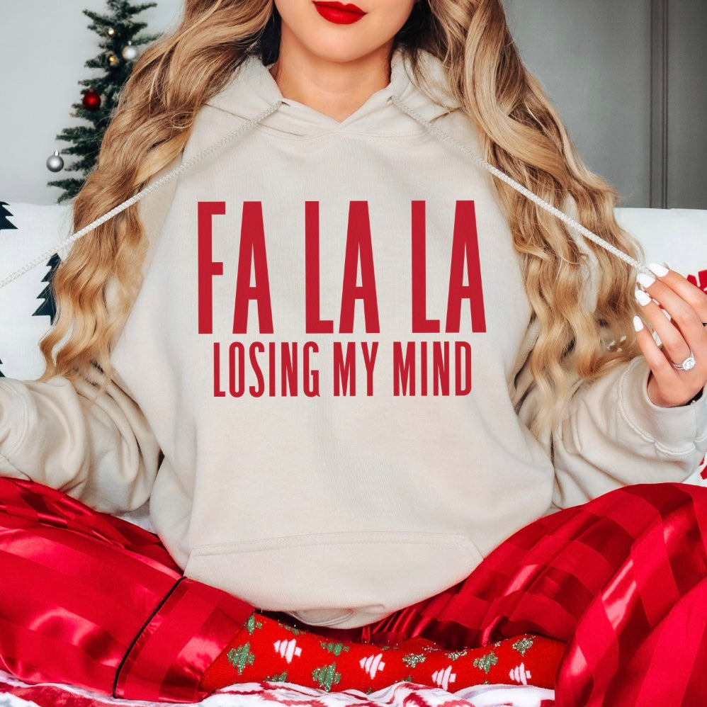A woman with long wavy hair sits cross-legged in red plaid pajamas, wearing the FA LA LA • Sand Hoodie featuring FA LA LA LA LOSING MY MIND in red text. A small Christmas tree is visible in the background.